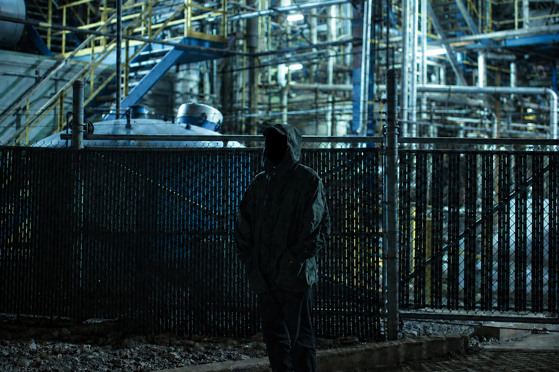 LXV with his face completely in shadow beside an illuminated industrial structure.