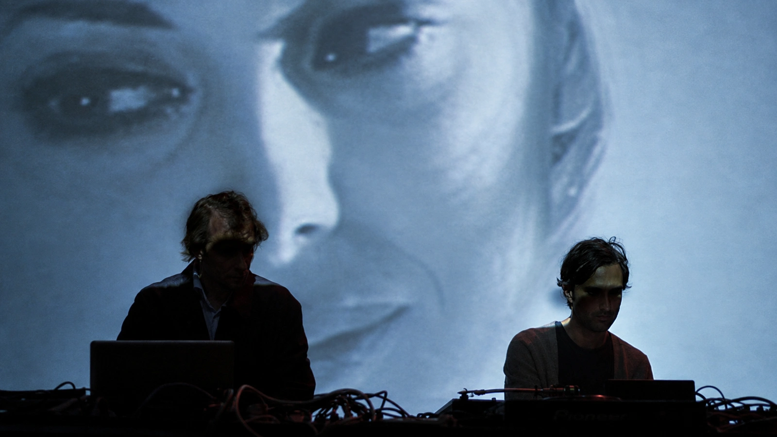 Demdike Stare performing in shadow beneath a blue-toned projection of a face.