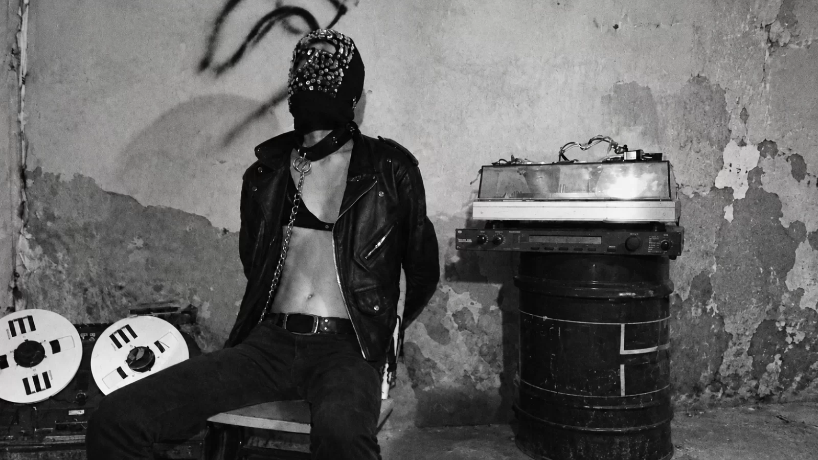 SARIN seated in a mask and leather jacket beside tape reels.