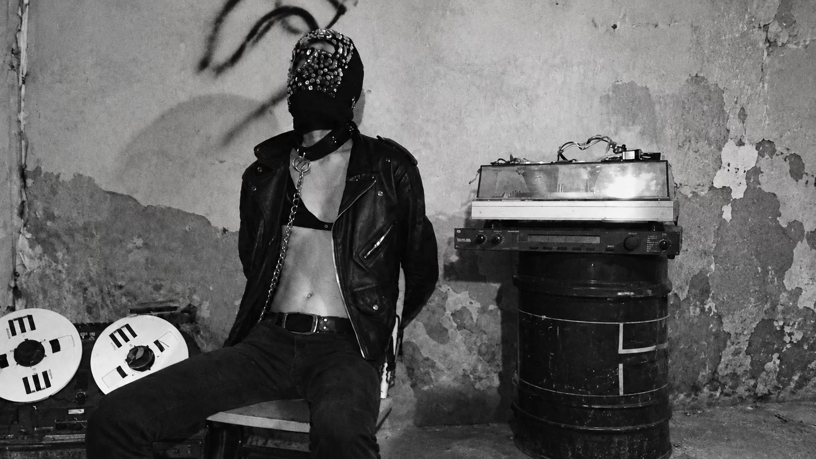 SARIN seated in a mask and leather jacket beside tape reels.