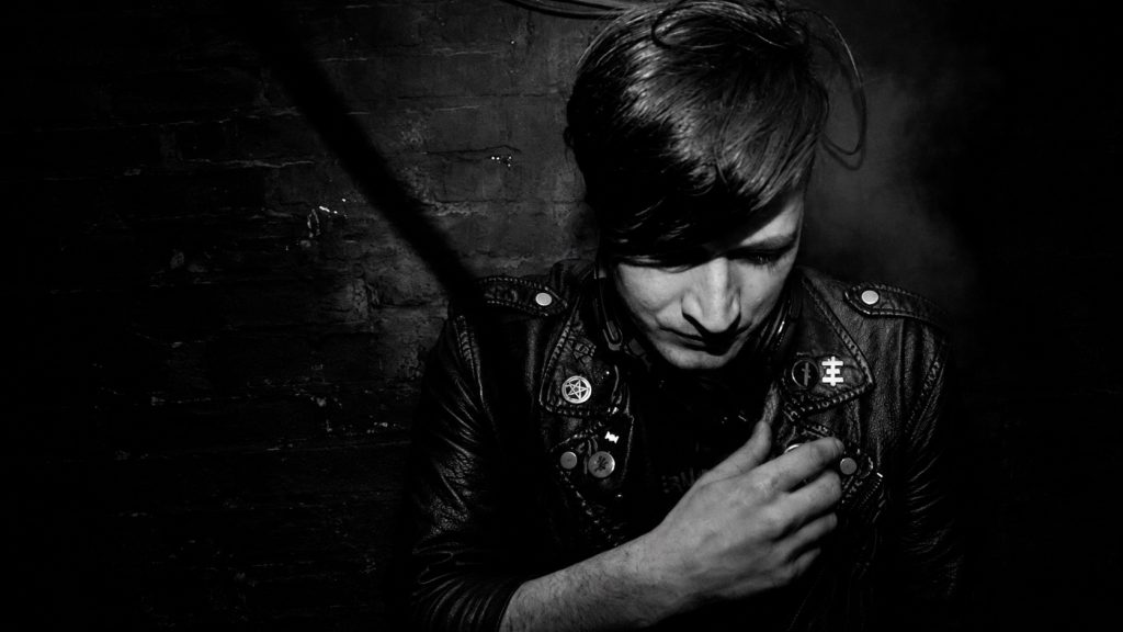 Philipp Strobel looking down in a leather jacket against a dark brick wall.
