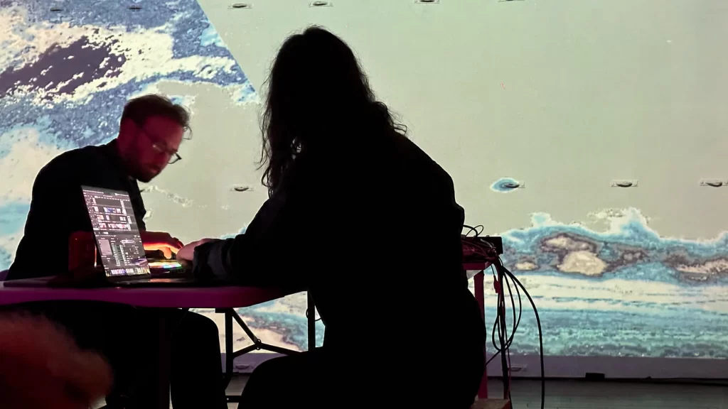 Emra Grid performing with another musician at a table of electronic equipment, with a projection behind them