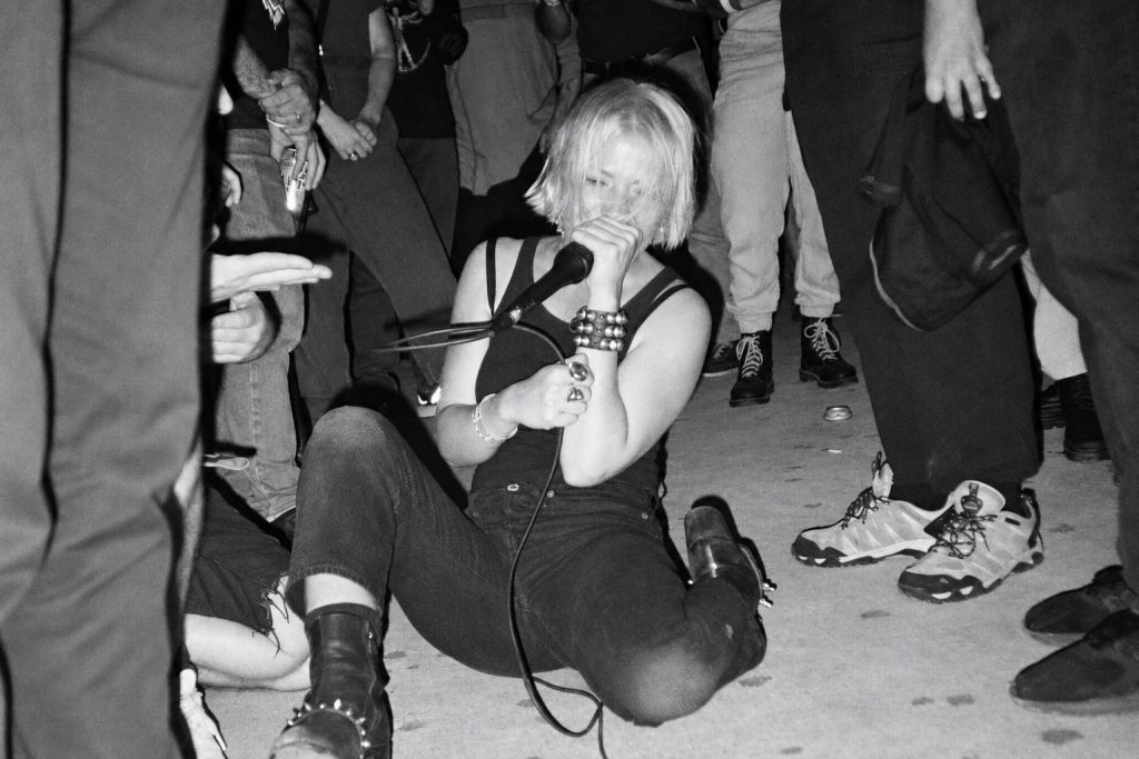 Pharmakon performing seated among the audience with a wired microphone