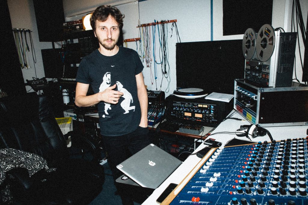 Lucy standing in his home studio beside electronic instruments and a reel-to-reel recorder