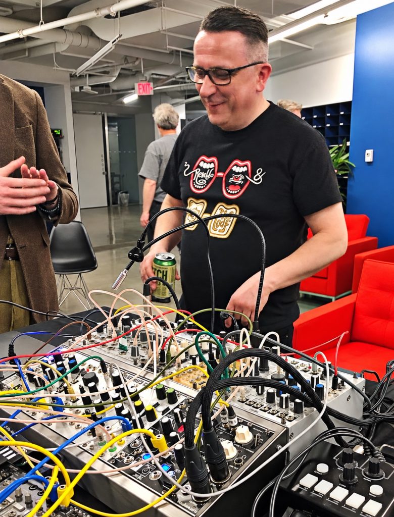 Peter Rehberg smiling beside a modular synthesizer in Boston, wearing glasses and a black graphic T-shirt.