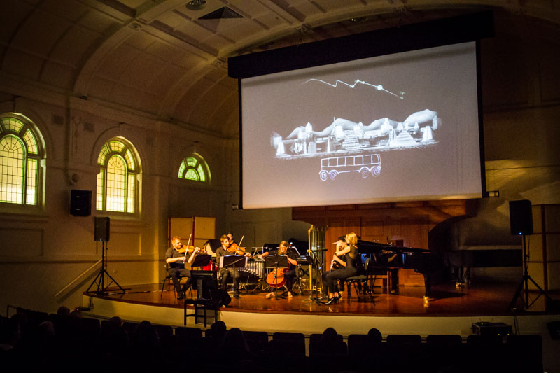 Rubiks Collective performing beneath projected drawings in Melba Hall, with a seated audience in front of the stage.