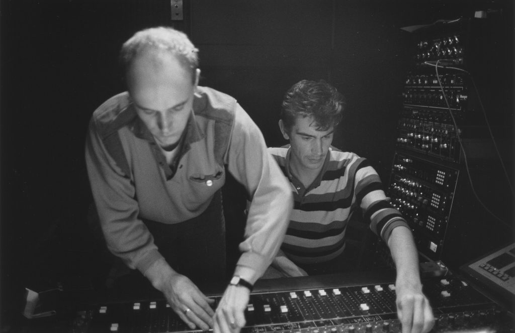 Mark Stewart and Adrian Sherwood leaning over a mixing desk in a recording studio.