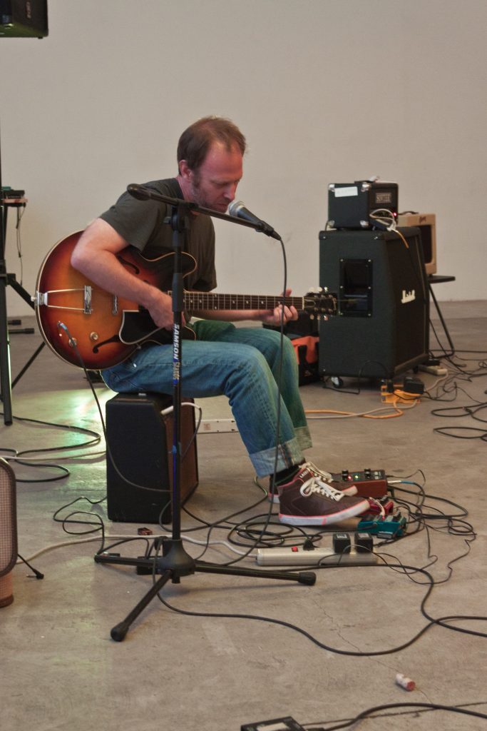 Steve Roden seated with a guitar and microphone during a performance at Human Resources in Los Angeles.